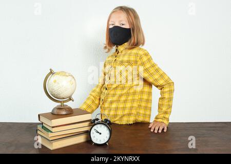 Concept d'éducation. Une fille dans un masque se tient près de la table. Il y a un globe sur la table, des livres et un réveil Banque D'Images