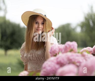 Jeune modèle posant dans un chapeau de paille à côté d'un grand buisson d'hortensia sur la toile de fond des arbres Banque D'Images