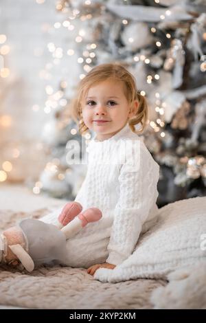 Très agréable charmante petite fille blonde en robe blanche assise sur un lit d'enfant et rires sur le fond des arbres de Noël dans l'intérieur lumineux de Banque D'Images