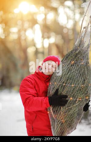 homme souriant portant un arbre de noël fraîchement coupé dans la forêt. Le jeune bûcheron porte un sapin sur son épaule dans les bois. Comportement irresponsable t Banque D'Images