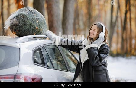 Happy Woman est en train d'écouter de la musique dans des écouteurs, de danser tout en emballant l'arbre de Noël avec le filet et les guirlandes sur un toit de sa voiture, se préparer à un Banque D'Images