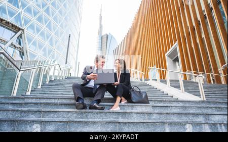 Un couple d'affaires travaillant sur un ordinateur tout en étant assis sur les escaliers des bâtiments financiers comme arrière-plan - deux hommes d'affaires analysant les finances Banque D'Images