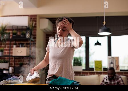 Femme souffrant de maux de tête sévères repassant des vêtements tandis que mari se détend sur le canapé à l'aide d'un téléphone portable. Femme épuisée finissant inconfortablement des tâches ménagères. Banque D'Images