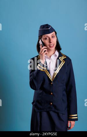 Préposé de vol souriant parlant sur smartphone, tenant le téléphone mobile, ayant la conversation. Hôtesse en uniforme répondant à un appel, hôtesse de l'air debout avec téléphone à la main, regardant la caméra Banque D'Images