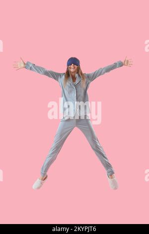 Jeune femme en pyjama et masque de sommeil sautant sur fond rose Banque D'Images