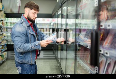 Un gars pensif à barbe qui fait des achats en magasin, en choisissant des saucisses fraîches pour le dîner Banque D'Images