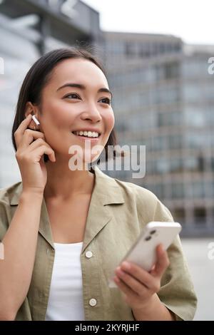 Prise de vue verticale d'une femme asiatique souriante dans un casque sans fil, aime écouter de la musique dans un casque, tient le téléphone portable Banque D'Images