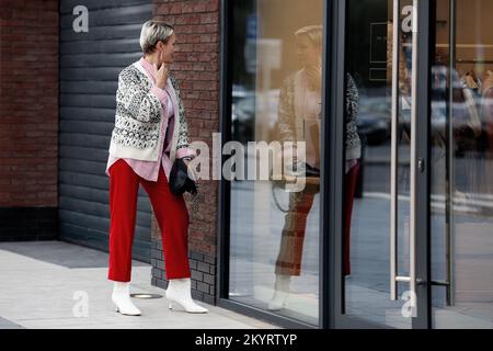 Femme habillée de façon tendance, regardant la réflexion dans le miroir de la fenêtre de magasin à l'extérieur. Modèle pour adulte avec chandail à cheveux courts et pantalon rouge. Banque D'Images