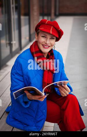 Femme élégante et à la mode, qui regarde un magazine et sourit à l'appareil photo. Modèle portant une veste bleue, un pantalon rouge et des accessoires vibrants béret ou chapeau, écharpe Banque D'Images