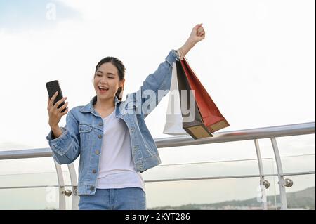 Une jeune femme asiatique joyeuse et heureuse se présente en avion, porte ses sacs d'achats et utilise son smartphone. Banque D'Images