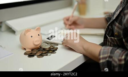 Gros plan, Banque Piggy et pièces de monnaie sur la table avec des femmes gérant et réalisant des comptes de revenus et de dépenses mensuels. Banque D'Images
