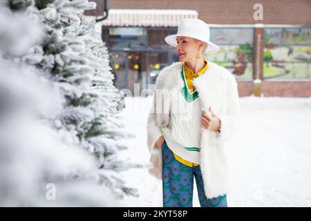 Hiver mode lumineux style de rue de la femme d'âge moyen, portant chapeau fedora et vêtements de manteau blanc en fausse fourrure avec des détails colorés Banque D'Images