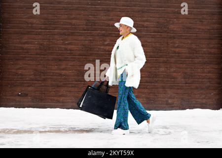 Mature heureuse femme tendance marcher dans la rue d'hiver avec des sacs de shopping, des cheveux courts de femme, portant chapeau feutre et des vêtements élégants et décontractés Banque D'Images
