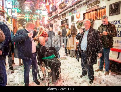 Fêtards qui chantent le long des Pogues, conte de fées de New York, alors que Guinness a donné vie à sa célèbre annonce de Noël à Belfast avec une machine à neige sur les rues pavées de commercial court devant le bar Duke of York, pendant que les invités ont été accueillis à une pinte pour commencer la saison des fêtes. Banque D'Images