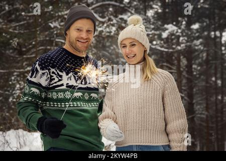 couple souriant avec des spameurs en plein air dans une forêt enneigée d'hiver. personnes avec des lumières du bengale brûlantes Banque D'Images