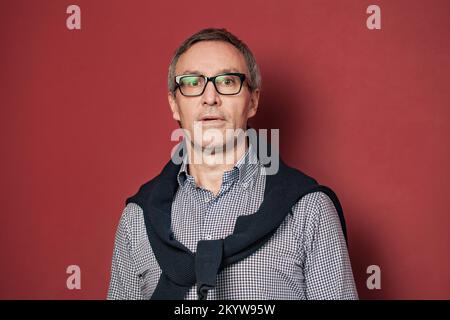 Portrait d'homme d'affaires sérieux et mûr à cheveux gris en chemise et lunettes sur fond rouge Banque D'Images