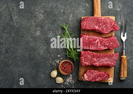 Steaks crus. Sur une planche à bois, placer les steaks sur la lame avec des épices, du romarin, des légumes et des ingrédients pour cuisiner sur fond noir. Vue de dessus. Copier