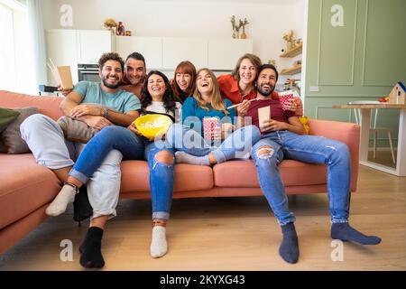 Un groupe d'étudiants qui regardent la télévision et qui mangent des en-cas assis sur le canapé - des jeunes étudiants gaies qui s'amusent dans le salon Banque D'Images