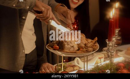 Gros plan d'un homme afro-américain coupant de la dinde ou du poulet. Famille multiculturelle célébrant Noël ou le jour de Thanksgiving. Table avec plats et bougies. Dîner de Noël familial à la maison. Banque D'Images