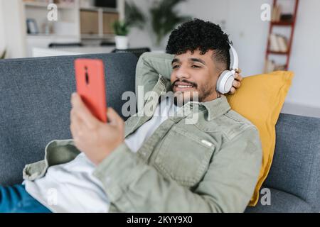 Un jeune homme latino-américain se détend sur un canapé en regardant les médias sociaux sur un téléphone portable Banque D'Images