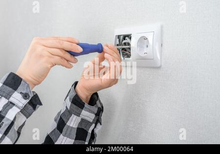 L'électricien utilise un tournevis pour installer la prise d'alimentation dans la boîte en plastique sur un mur blanc. travail dangereux avec un réseau électrique haute tension. Main wi Banque D'Images