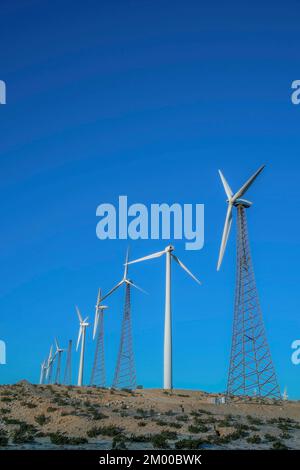 Éoliennes sur une pente de montagne dans un désert en Californie. Moulins à vent avec des tours tubulaires en acier et en treillis contre le fond bleu clair du ciel. Banque D'Images
