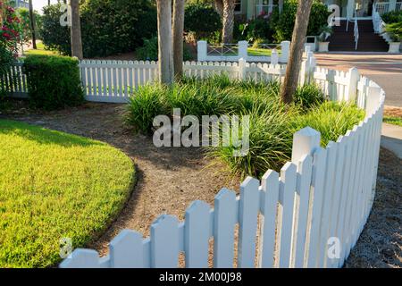 Destin, Floride - clôture de piquetage blanche peinte avec coin incurvé. Il y a de l'herbe tondue à gauche près des buissons et des arbres à côté de la clôture. Banque D'Images