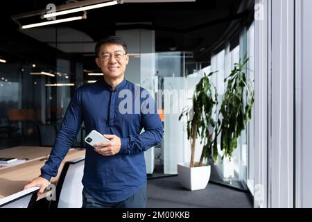 Portrait d'un homme d'affaires asiatique réussi, homme en lunettes et chemise souriant et regardant l'appareil photo, investisseur réussi tenant le téléphone debout près de la fenêtre. Banque D'Images