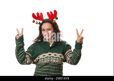 Jolie femme en chandail à motifs de Noël vert et cerceau de cerf, montrant le signe de paix sur l'appareil photo sur fond blanc Banque D'Images