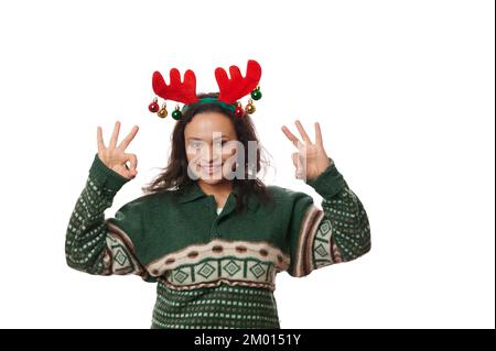 Belle femme en vert chandail à motifs de Noël et cerceau de cerf, montrant le signe OK sur l'appareil photo sur fond blanc Banque D'Images