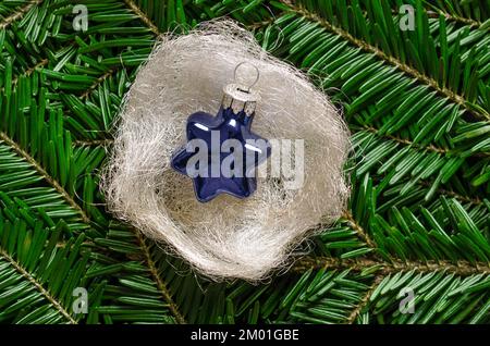 Étoile en verre bleu, dans un nid de cheveux d'ange blanc argenté, sur des branches de sapin vert frais. Boule en verre en forme d'étoile à suspendre sur un arbre de Noël. Banque D'Images