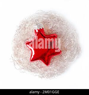 Étoile en verre rouge, ancrée dans un nid de cheveux d'ange blanc argenté, sur un fond blanc. Une boule en verre en forme d'étoile, à suspendre sur un arbre de Noël. Banque D'Images