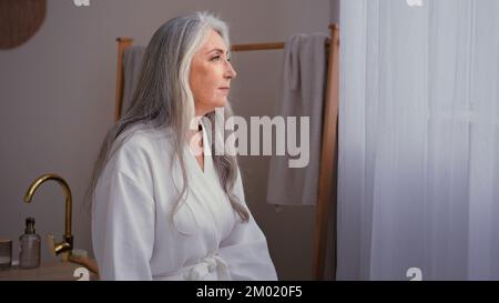 Femme âgée pensive 60s ans caucasien 50s senior mature magnifique femme en blanc peignoir debout dans la salle de bains à la maison. Calme grand-mère femme Banque D'Images