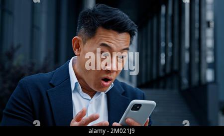 Heureux choqué 50s ans d'homme en costume formel debout en plein air regardant le téléphone dans la ville 40s gagnant homme d'affaires a le succès d'affaires des nouvelles étonnantes Banque D'Images