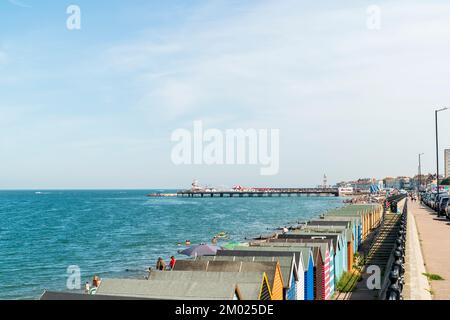 Bord de mer de Herne Bay lors d'une chaude journée d'été. Rangée de cabanes de plage, voitures garées, avec la plage animée, la jetée et tour d'horloge dans la distance sous un bleu s Banque D'Images