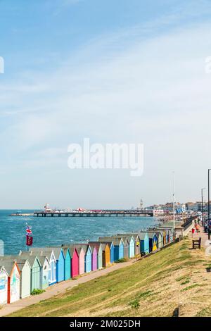 Bord de mer de Herne Bay lors d'une chaude journée d'été. Rangée de cabanes de plage, avec la plage animée, la jetée et la tour de l'horloge au loin sous un ciel bleu. Banque D'Images