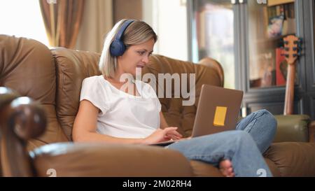 Femme indépendante avec casque et ordinateur portable travaille en ligne tout en étant assise sur un canapé. Banque D'Images