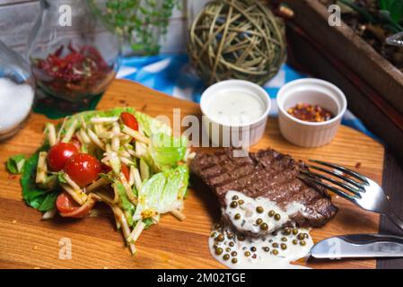 Un morceau de bœuf grillé à la sauce au poivre vert sur une planche de bois avec une salade de pommes, de tomates cerises et de feuilles de laitue iceberg Banque D'Images