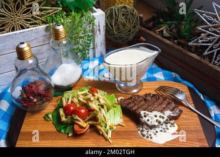 Un morceau de bœuf grillé à la sauce au poivre vert sur une planche de bois avec une salade de pommes, de tomates cerises et de feuilles de laitue iceberg Banque D'Images