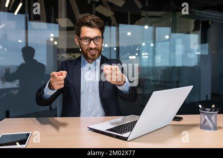 Homme d'affaires mature et réussi à regarder la caméra et de pointer du doigt vers l'avant appelant à l'action, investisseur en costume d'affaires dans le bureau moderne travaillant sur ordinateur portable, patron avec la barbe et les lunettes. Banque D'Images