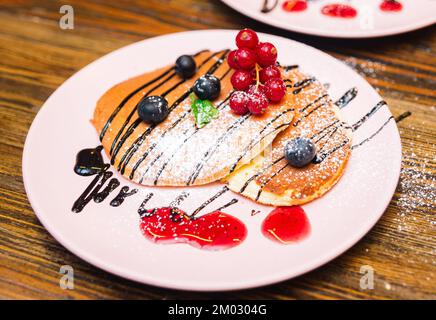 Assiette rose avec crêpes soufflées aux raisins de Corinthe, bleuets, saupoudrés de sucre en poudre, sauce au chocolat Banque D'Images