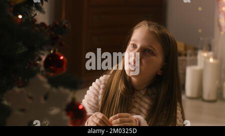 Une jeune fille en pyjama doux est assise sur le sol, décorera l'arbre de Noël avec des balles et des jouets, sourit et passe du temps libre à la maison. L'enfant prépare sa chambre avant les vacances d'hiver et le nouvel an. Ralenti. Banque D'Images