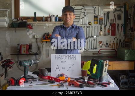 Image d'un homme souriant dans son atelier derrière un banc rempli d'outils de travail avec un homme de a à Z écrit dessus. Homme qui peut tout réparer Banque D'Images