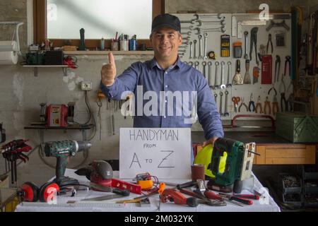 Image d'un homme souriant dans son atelier avec un pouce derrière un banc rempli d'outils de travail avec un homme de a à Z écrit dessus. Homme qui peut fixer et Banque D'Images