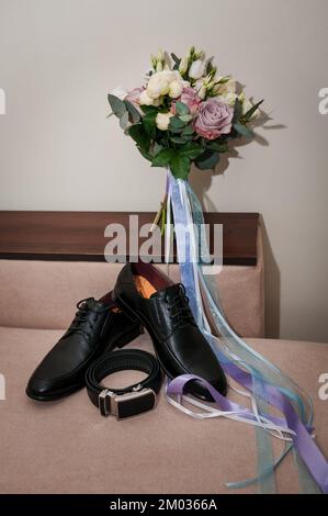 Accessoires pour marié. Chaussures en cuir noir pour hommes, bouquet de mariée, ceinture en cuir noir sur fond marron. Banque D'Images