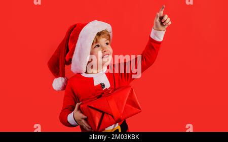 Aide du père Noël. Petit enfant garçon dans un chapeau de Noël avec boîte cadeau actuelle pointant vers l'espace de copie. Banque D'Images