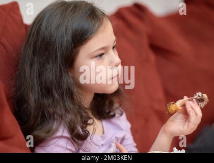 La jeune fille expressive mange du poulet et des légumes pour le dîner sur le canapé tout en regardant la télévision Banque D'Images