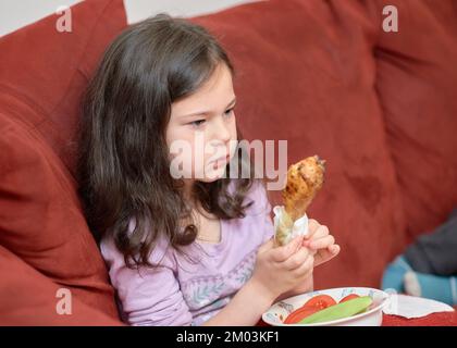 La jeune fille expressive mange du poulet et des légumes pour le dîner sur le canapé tout en regardant la télévision Banque D'Images