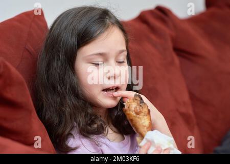 La jeune fille expressive mange du poulet et des légumes pour le dîner sur le canapé tout en regardant la télévision Banque D'Images