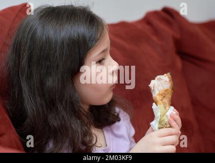 La jeune fille expressive mange du poulet et des légumes pour le dîner sur le canapé tout en regardant la télévision Banque D'Images